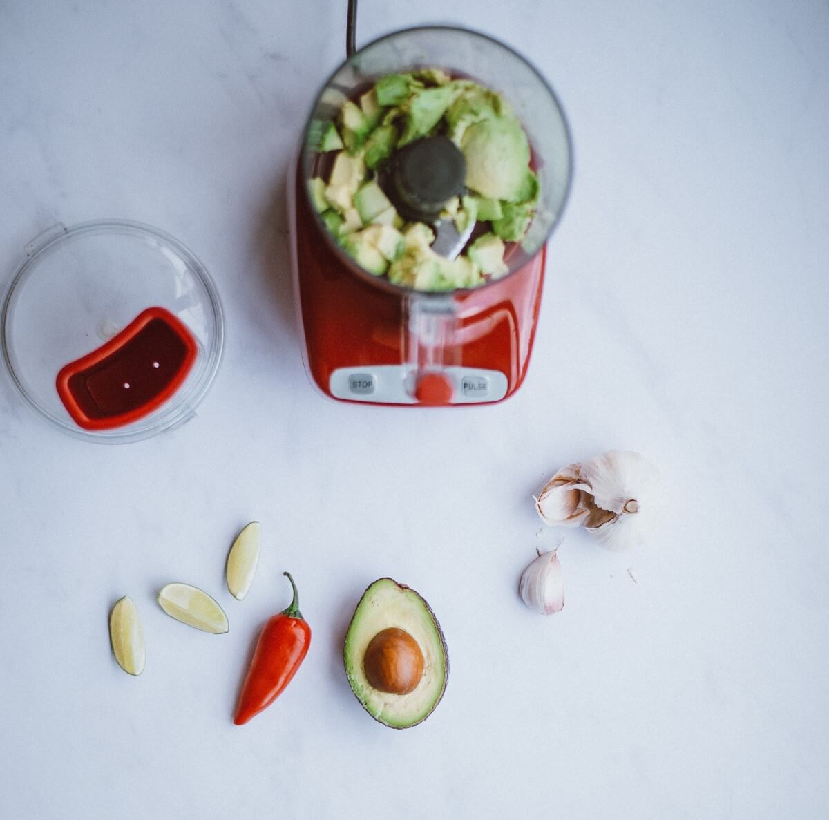 Mixer auf hellblauem Hintergrund, Im Mixer eine leicht pürierte Avocado. Auf dem Tisch liegt eine Waage, Knoblauch, eine Chilichote und eine halbe Avocado mit Kern