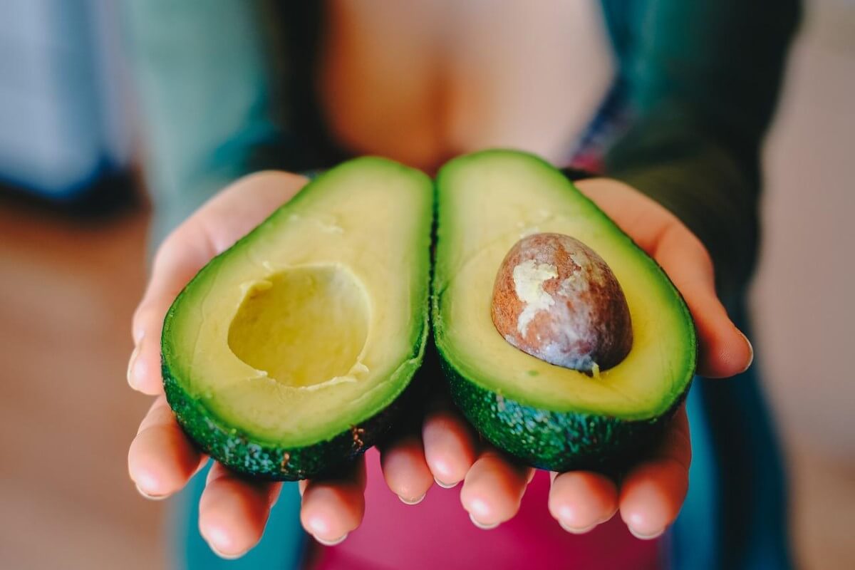 Zwei Avocadohälften in Menschenhänden. In der rechten Avocadohälfte liegt der Kern