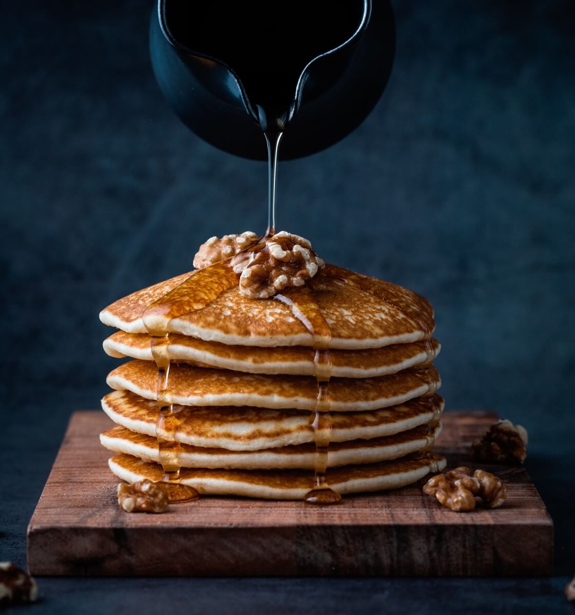 Pancakes auf einer Holzplatte. Garniert mit ein paar Walnüssen. Mit einer schwarzen Kanne wird Agavendicksaft darübergeschüttet