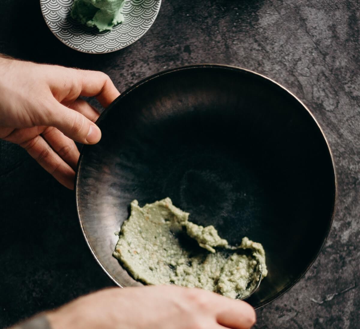 Wasabimayonnaise in einer schwarzen SChale auf Betonfarbener Arbeitsplatte. Im Hintergrund Wasabi auf einem hellgrauen kleinem Teller. Zwei Hände rühren mit einem kleinen Löffel in der Wasabimayonnaise