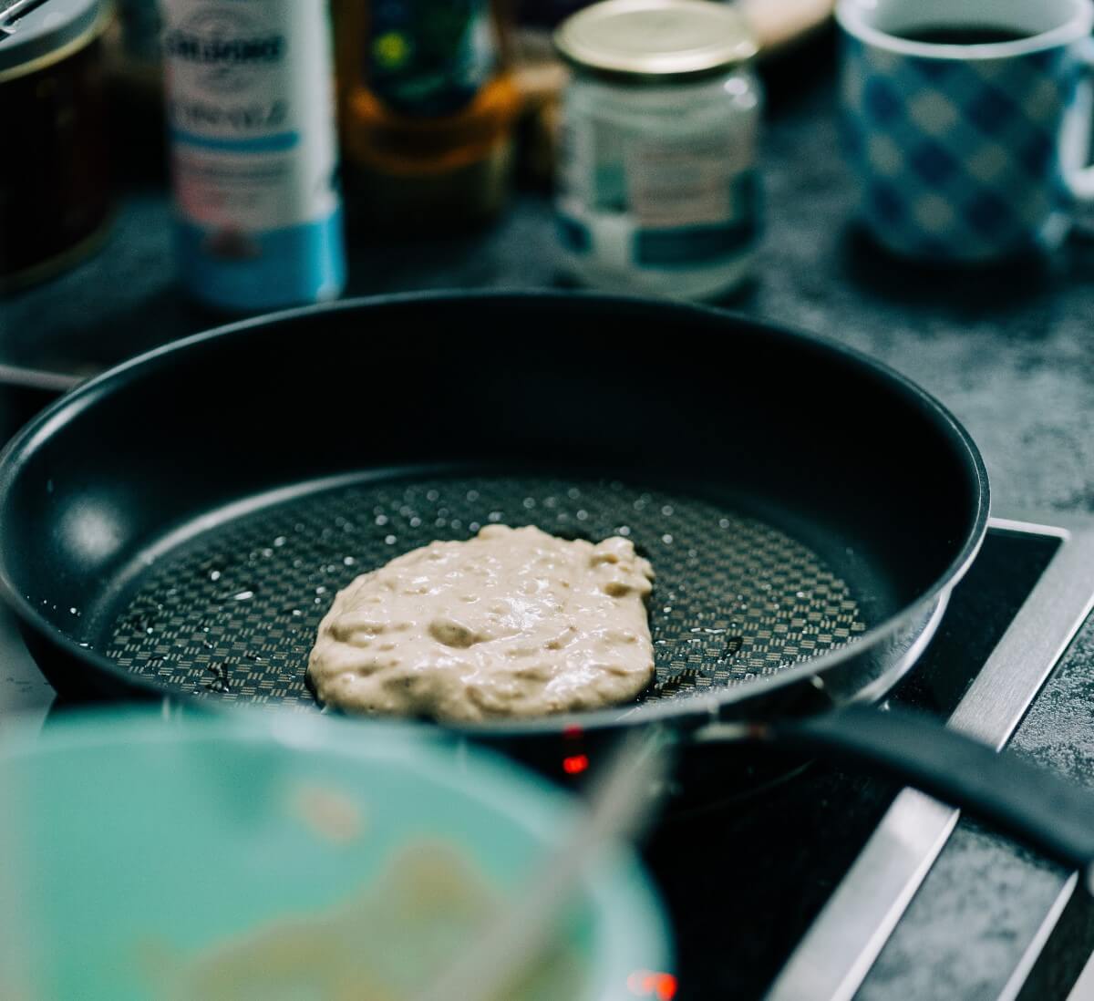 Pancake-Teig in einer Pfanne auf dem Ceranfeld. Auf der Arbeitsfläche stehen einige Behälter